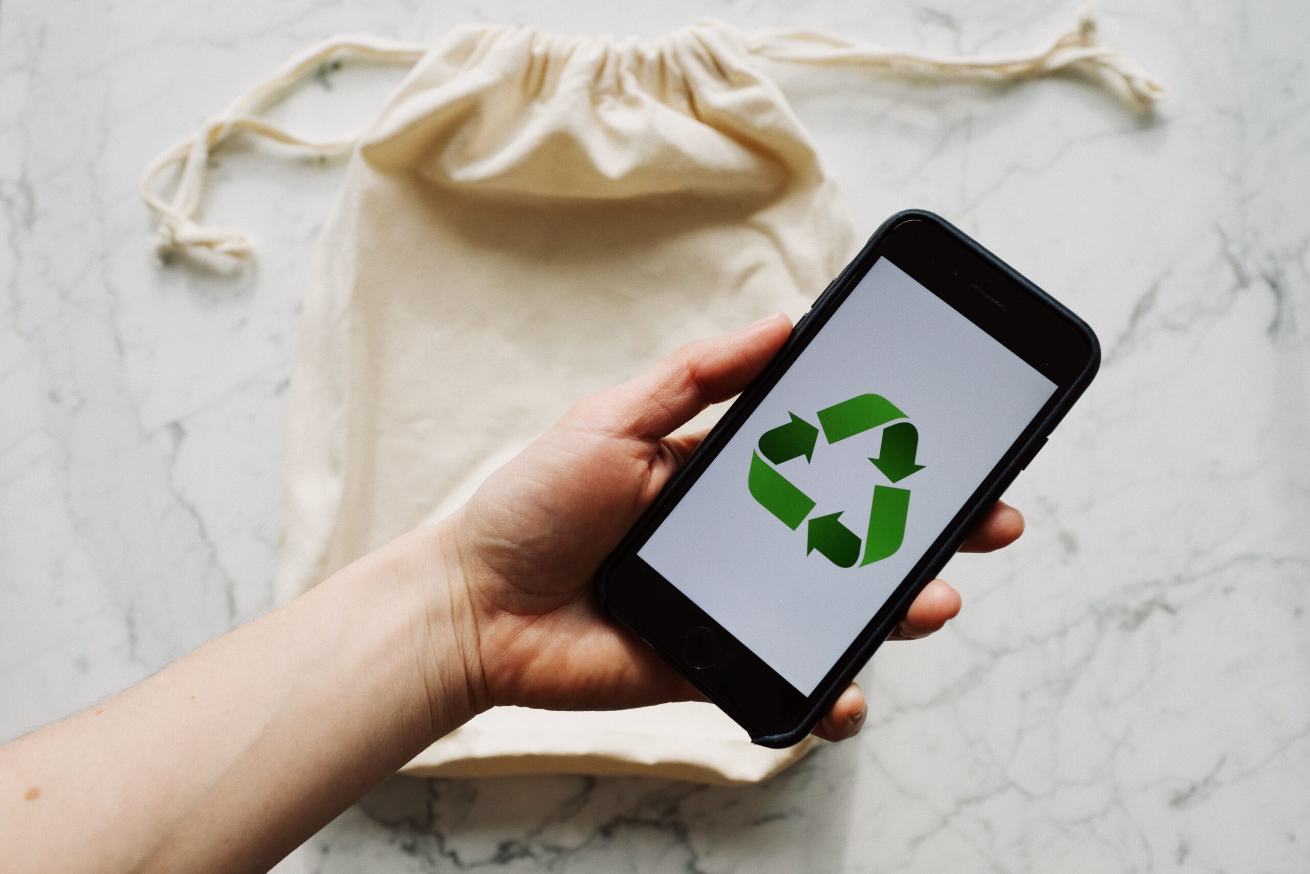 From above view of faceless person holding smartphone with green recycling symbol on screen and white textile bag placed on marble table