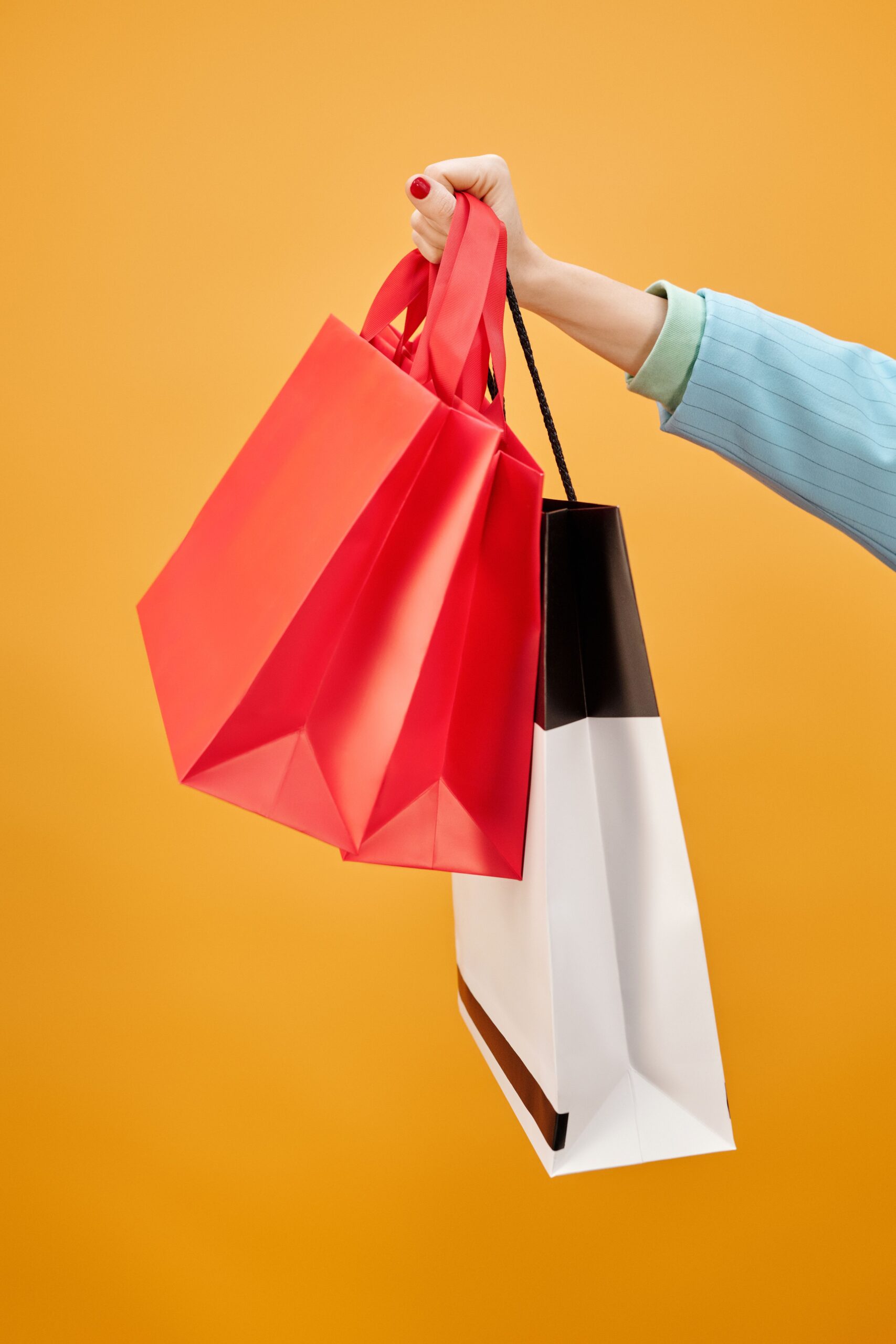 A hand holding vibrant shopping bags on a bright yellow background, symbolizing retail and consumerism.